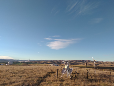 photo of eddy covariance system at a bit of a distance with pasture behind, research facility in the background with partly cloudy sky